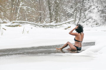 Beautiful girl sitting on the ice the cold water of a lake or river, cold therapy, ice swim with winter landscape and forest on background
