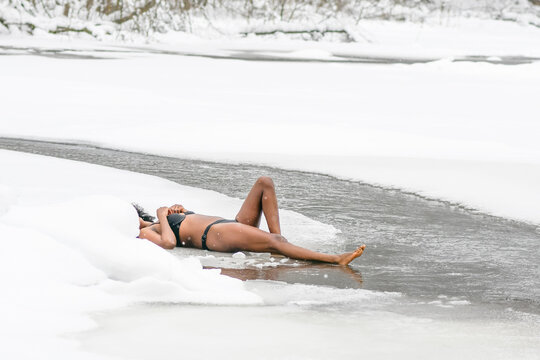 Beautiful girl lying on the ice near cold water of a lake or river, cold therapy, ice swim with winter landscape and forest on background