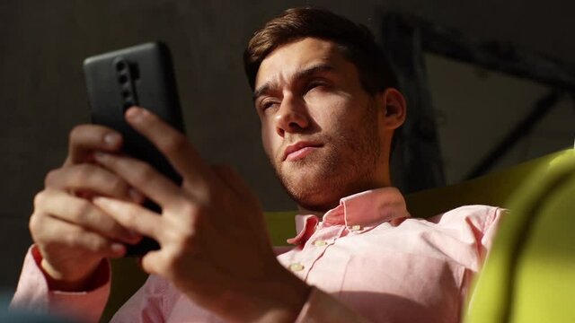 Close up of tired young man having blurry vision is rubbing and squinting eyes while typing using smartphone, sitting in yellow armchair in dark room. Bored exhausted male checking mobile phone.