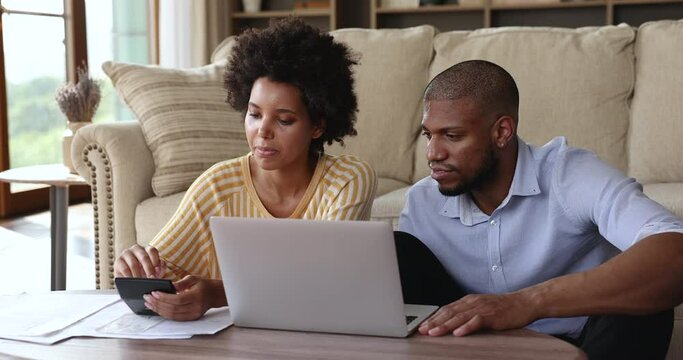 Focused Afro American spouses sit by laptop at home office calculate bills to pay online manage family finances engaged in budgeting work. Young couple prepare loan mortgage payments plan investment