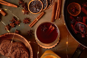 glass of hot mulled wine with stick of cinnamon and star anise between saucepan where brewed wine, dried citrus fruits, jar of honey and ingredients, wooden bowl with gingerbread and glowing garland