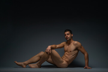 Gorgeous naked male model sitting isolated on the floor in a studio while looking in the camera