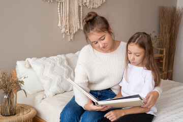 mother and daughter are sitting at home on the bed flipping through and looking at a book with photos. important moments of life in a photo album. Cute sisters read an interesting fairy tale together