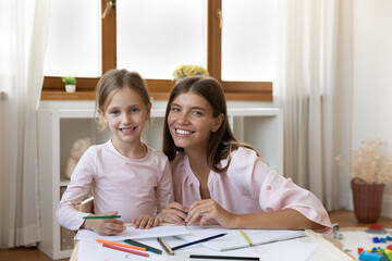 Female art teacher and little learner portrait concept. Loving young mother her cute preschool 6s daughter smile look at camera, enjoy hobby at home, use colour pencils drawing pictures in sketchbook