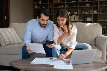 Focused husband and wife sit at table at home pay utilities online, young couple make internet...