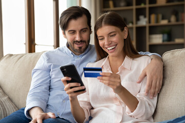 Beautiful couple sitting on couch using phone and plastic card buy goods and services on internet,...
