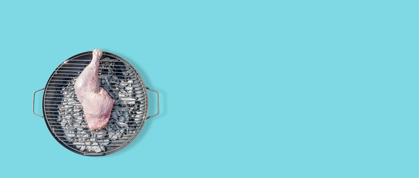 Banner with big chicken leg prepared for grill at a round barbeque firepan isolated at turquoise solid background with copy space. Concept of food in outdoor cooking.