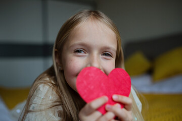 Little school kid girl holding red heart. Valentine's day during pandemic coronavirus covid-19 quarantine. Child showing heart online via video chat to boy friend
