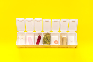 White pillbox with a big cannabis bud and different colorful pills on yellow background. Concept of medical marijuana and CBD, natural medicines...