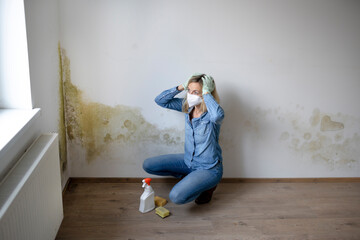 blonde young woman sitting in front of white apartment wall with mold on it frustrated and desperate
