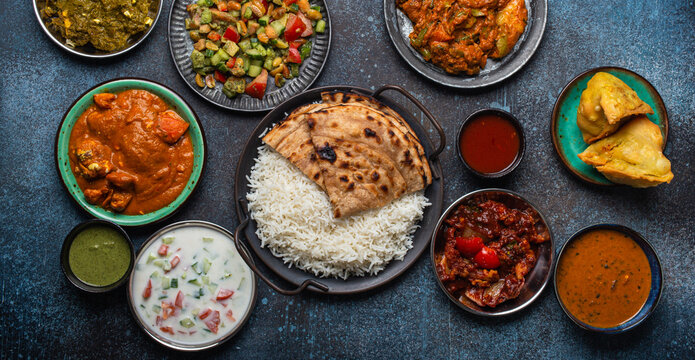 Assorted Indian ethnic food buffet on rustic concrete table from above: curry, fried samosa, rice biryani, dal, paneer, chapatti, naan, chicken tikka masala, mango lassi, dishes of India for dinner