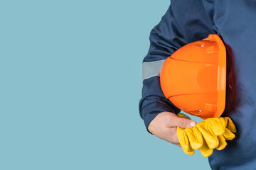 A man in work clothes holds an orange protective helmet and yellow gloves on a blue background in...