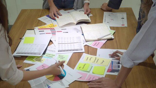 Upper view of many paper sheets with chart, data, report, sticky notes, and notebook about a work plans, scattered on a table with a group of business people's hands writing on papers during a meeting
