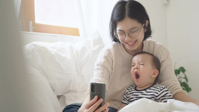 Smiling Chinese Young mother and her cute boy using smartphone at home, Asian family doing video call on mobile phone, Little son yawning