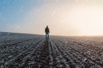 Young caucasian man in jacket standing on meadow, field covered with hoarfrost in misty fog at...