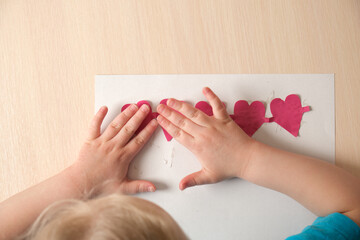 preschooler girl in a blue t-shirt makes an applique sitting at the table, hearts cut out of colored bougue for a card