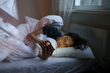 Social media jealousy. Young depressed woman addicted to mobile phone lying in bed at home,...