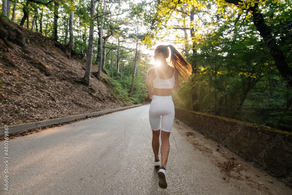 Wall mural a young beautiful girl in white sports clothes is running with her back, on the road in a dense fore