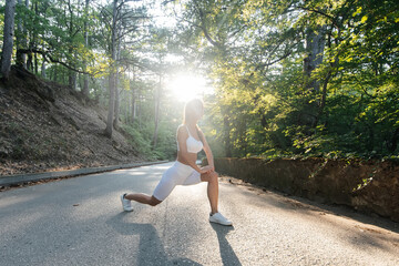 A young beautiful girl makes lunges and warm-up before running training, on the road in a dense...