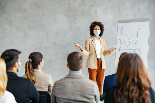 Broker presentation and seminar about the economy. An African American female broker with a face mask on her face stands in the boardroom next to a chart in front of the people and has a speech.