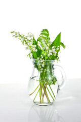 Lily of the valley or may bells flower with green leaves in a glass jug on a white background.