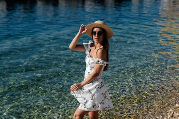 A beautiful young woman in a hat, glasses and a light dress is walking along the ocean shore against the background of huge rocks on a sunny day. Tourism and tourist trips.
