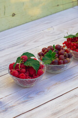 Raspberries, red currants, gooseberries, black currants in glass plates on a rustic white wooden table