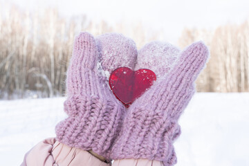 The concept of the holiday is Valentine's Day. Heart in hands in warm mittens amid winter forest