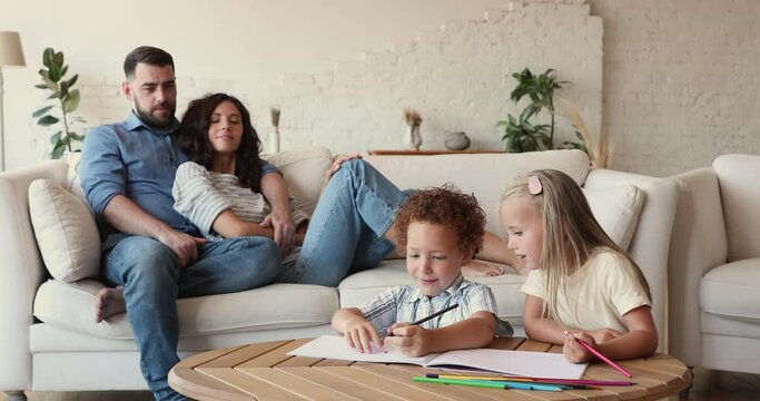 Multi ethnic family spend time on weekend together at modern home, preschool diverse children friends drawing with pencils on album, parents relaxing seated on cozy sofa enjoy leisure. Pastime concept