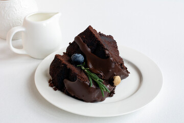 Chocolate cake with icing, decorated with fresh blueberry and green rosemary twig, on plate on table with sugar bowl and creamer  on light background