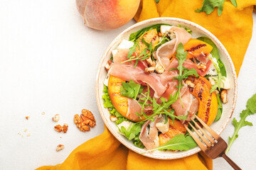 Salad bowl with grilled peach, jamon, feta cheese, nuts and arugula on white table background, top view, copy space