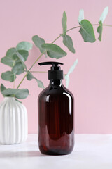 Dark amber glass shampoo dispenser bottle and eucalyptus in vase on pink background. Cosmetics product branding mockup.