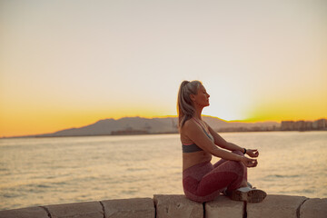 Fototapeta na wymiar Side view of sporty lady practicing yoga, sitting on edge at seaside on background of sky at sunset and mountains