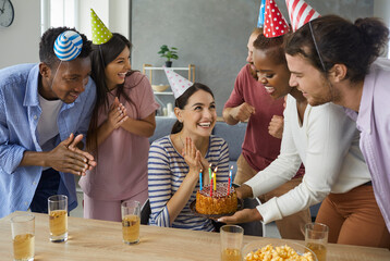 Fototapeta Diverse group of cheerful friends in their twenties giving a cake with burning candles to a happy birthday girl. Young woman in her 20s getting presents and having fun at her birthday party at home obraz