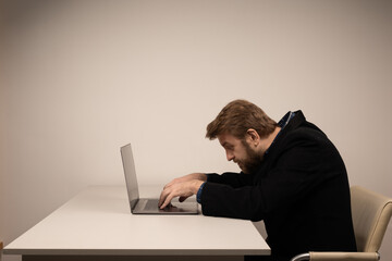 Beard emotional man in slouching position sitting in office room, working with laptop