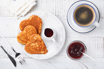 Pancakes with berry jam and honey in shape of heart and hot cup of coffee over white wooden background. Concept breakfast for Valentines Day or pleasant surprise for loved one Table viewed from above
