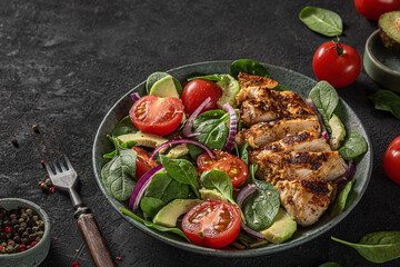 Grilled chicken breast fillet and spinach salad with avocado, tomatoes and sesame seeds in a plate on dark background