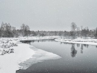 Winter landscape with small river in wood on background gray snowy sky.