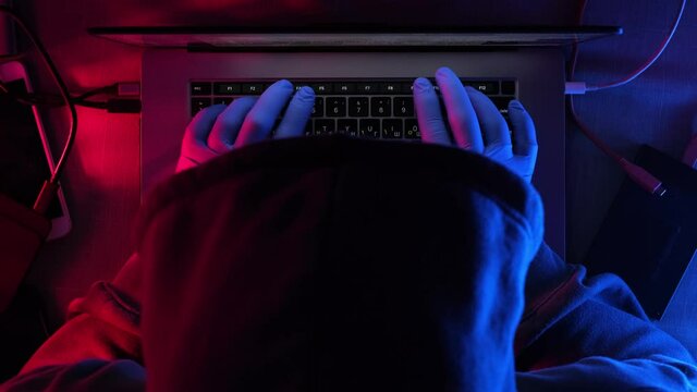 hacker works behind a laptop in a dark room. the room has red-blue light and the effect of the night. hacker in a hood and with gloves.
