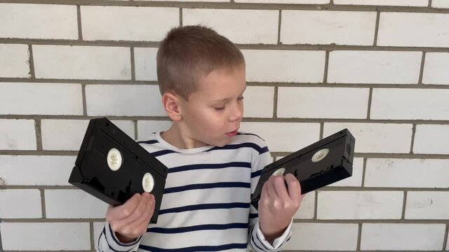 A shocked boy on the background of a brick wall is holding a videotape, a very popular technology of the 90s, and looks at the tape in surprise.