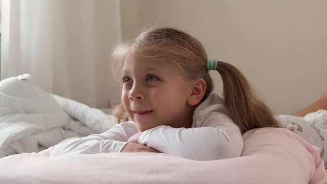 A beautiful little preschooler girl in white pajamas on the bed watches TV intently. The concept of children's addiction. Watch over the children. Day off, hobby. Children and TV Addiction
