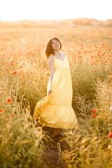 Beautiful young woman in a yellow dress walking in a poppy field on a summer day. Girl enjoying flowers in the countryside. Selective focus