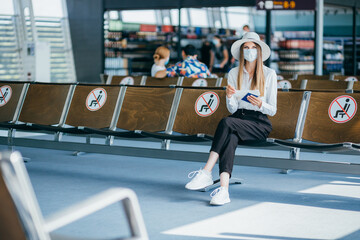 Young woman tourist wearing face mask holding passport and ticket, sitting in airport or train station on empty marked chairs under new normal and social covid distance regulations.