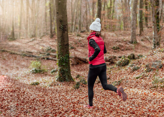 middle-aged woman in white woolen cap and pink vest, running through a forest on dry leaves at...