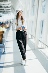 Elegant beautiful business girl holding travel tickets and using phone, happy girl looking at camera in airport terminal. Fashion woman portrait. Airport terminal. Business trip.