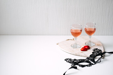 Valentines Day Festive table setting with black lace mask, two glasses of rose wine and two red heart shape candies on white table. Valentine Day, flirting, romance, dating concept, copy space