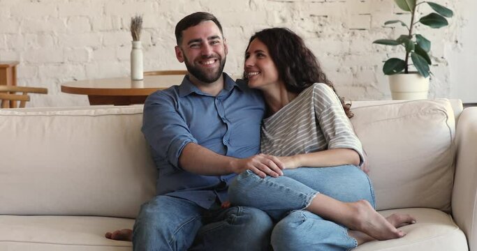 Wife and husband sit on sofa smile pose look at camera. Cheerful couple relaxing at home staring aside daydreaming enjoy weekend leisure at their own or rented house. Homeowner family portrait concept