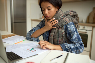 Sick African female student having cold and sore throat, sitting at kitchen table in front of...