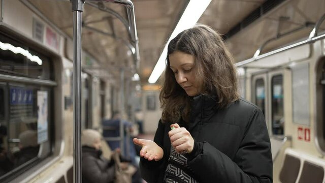 Young squeamish woman with dark wavy hair wearing warm jacket uses sanitizer on hands after holding handrail in underground carriage