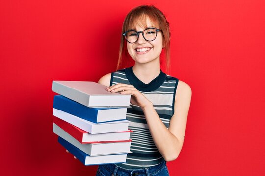 Redhead young woman holding a pile of books smiling with a happy and cool smile on face. showing teeth.